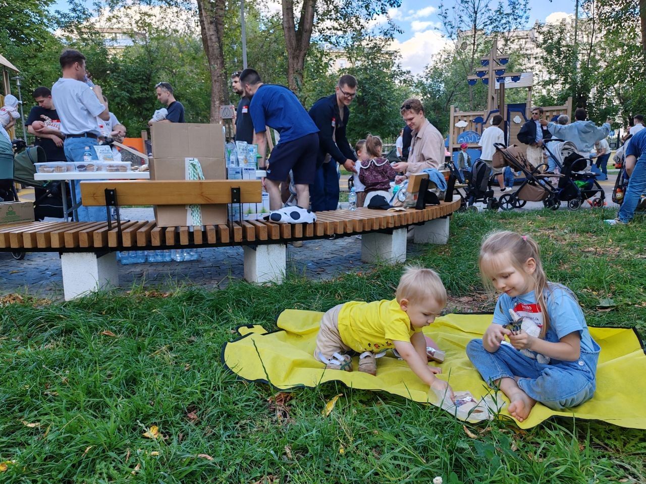 Двое малышей играют на жёлтом пледе на траве, участники встречи рядом