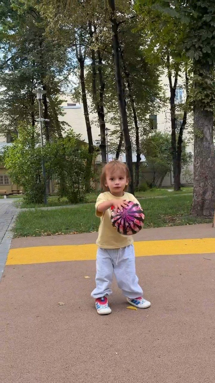 Малыш в жёлтой футболке держит мяч на дорожке сада